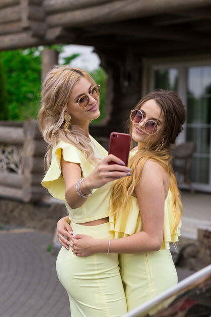 肖像两位穿着黄色礼服的年轻漂亮伴娘的画像 就在一个时髦的木制房间旁边房子人生活方式概念.行动手机自拍微笑送空吻微笑青春快乐