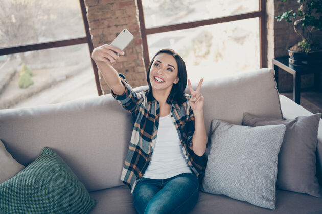 博客迷人女士手持电话为博客自拍的照片显示 v字符号坐在舒适的沙发上 穿着休闲格子衬衫和牛仔裤公寓室内酷模型房间