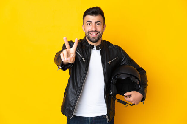 表情高加索男子戴着摩托车头盔 在孤立的黄色背景下微笑着 展示着胜利的标志摩托车皮革男性
