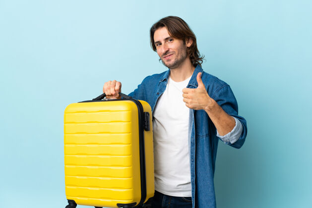 瘦身年轻英俊的男人在度假时被隔离在蓝色的墙上 提着旅行箱 竖起大拇指公文包拇指行李