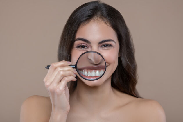 牙齿健康的牙齿年轻快乐的女人 光着肩膀 在微笑的嘴边拿着放大镜 露出大大的白牙裸露的肩膀看深色头发