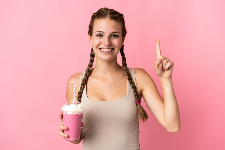 年轻人一个年轻的女人用草莓奶昔隔离在粉红色的背景指向一个伟大的想法微笑演示水果