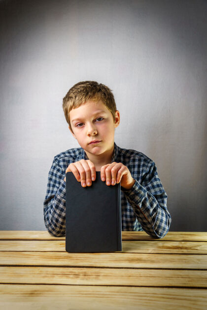 读者悲伤的男孩从7岁到9岁 手里拿着一本书 背景是灰色的一张木桌教育小学生聪明