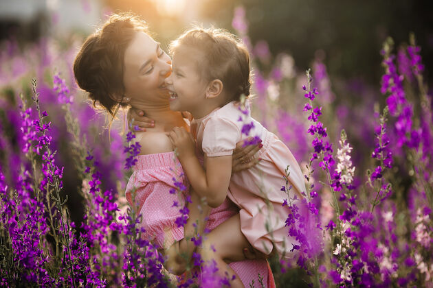 欢呼年轻漂亮的妈妈和她的小女儿一起在户外玩耍花紫罗兰色花坛成人关怀日落