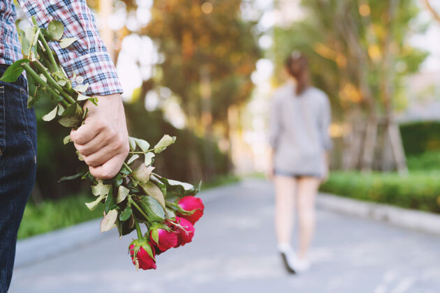 微笑年轻人手拿红玫瑰站在悲伤的爱情中结束了分手的恋情迷离的女人背影在公园里走远了在户外分手破碎的心在情人节的概念空间爱花