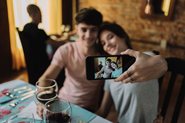 姿势一对微笑的年轻夫妇坐在餐厅里 喝着酒——一对快乐的年轻夫妇用智能手机自拍手图片眼镜