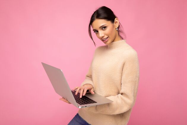 空闲时间这张照片是一位身穿米色毛衣的年轻黑发美女手持上网本 在键盘上输入文字 看着隔离在粉色墙壁背景上的相机学生工作学习