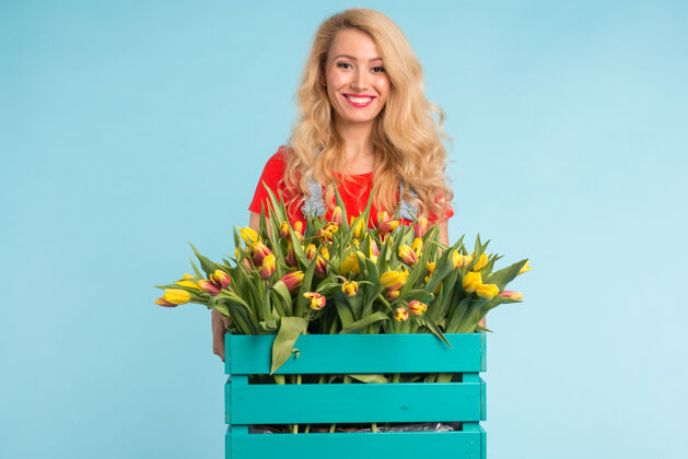 花美丽的金发女园丁拿着盒子 蓝色的表面上有郁金香花束礼物