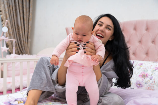 快乐妈妈扶着宝宝站着 抱着她的胸脯 坐在铺着粉色床单的床上 看着相机笑着人女儿在一起