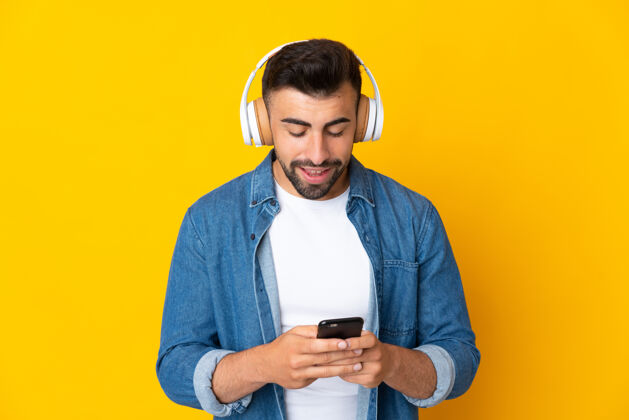 快乐白人男子在孤立的黄色背景下听着音乐 看着手机西班牙裔拉丁语耳机