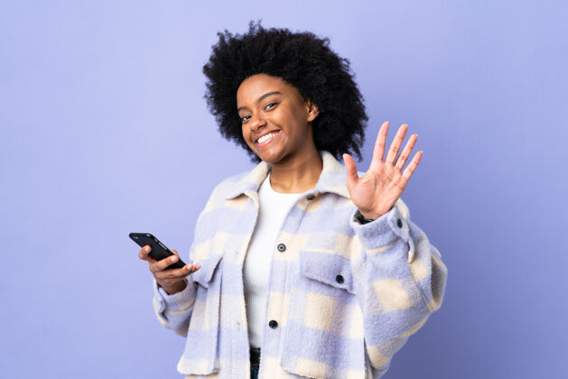 美国人年轻的非洲裔美国妇女用手机孤立地用紫色的手敬礼 表情愉快通讯朋友人