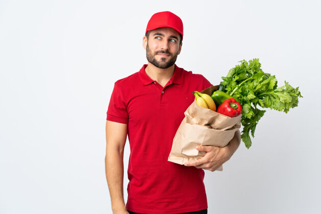胡须留着胡子的年轻人拿着一个装满蔬菜的袋子 孤立地站在白色的背景上 望着旁边外卖生的健康