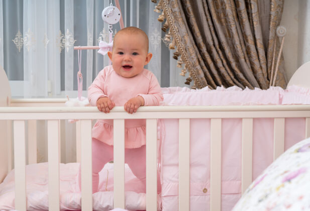 警觉一个快乐的 笑着的婴儿站在婴儿床上的粉红色卧室场景复制空间移动的新生儿