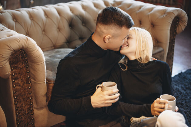 男性一个迷人的年轻人和女人坐在家里的地板上喝着热咖啡 一边笑着玩的侧视图关系拥抱圣诞节