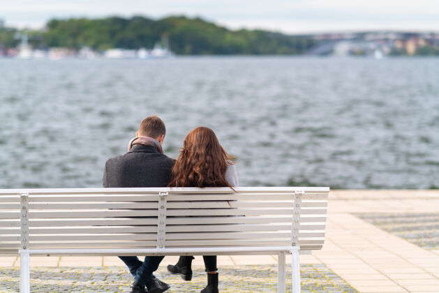放松秋风凛冽的日子里 一对穿着厚外套的夫妇坐在长凳上俯瞰着波涛汹涌的水面 从后视图中可以看到朋友观光男朋友