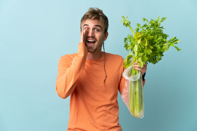 生的年轻的金发男子拿着一个芹菜孤立在蓝色的叫喊与张大嘴巴青年蔬菜男性