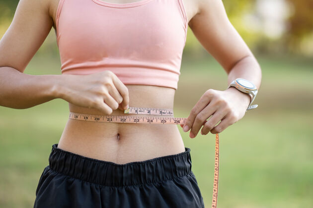 体重年轻苗条的女性 穿着粉色运动服 健康的女人在公园里用卷尺测量她纤细的腰室外重量减肥 健身 运动理念磁带大小运动