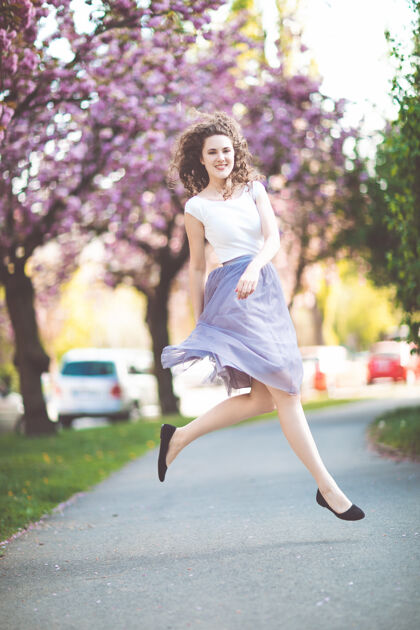 成人一头卷发 身穿白色t恤和紫罗兰色裙子的年轻女孩在盛开的樱花树下翩翩起舞肖像年轻积极