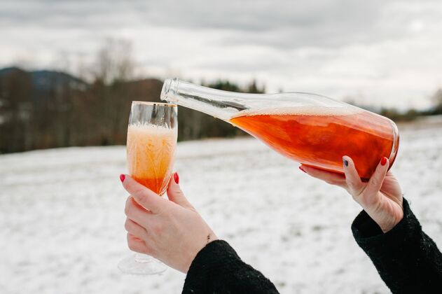 酒女人手里拿着一瓶香槟 倒进了一个以冬山为背景的玻璃杯里雪女性香槟