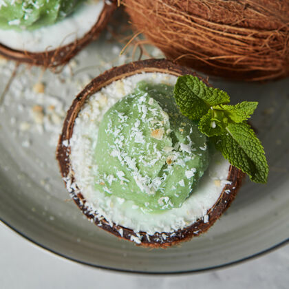 吃自制的绿色冰激凌在椰子壳里 薄荷叶放在灰色的陶瓷盘上灰色上衣查看素食者饮食的概念干净椰子素食