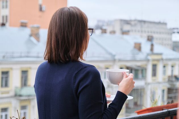 房子成熟的女人端着一杯茶站在开放的阳台上 女人一边看城市一边隔离 隔离期间病毒感染 复制空间看窗户污染