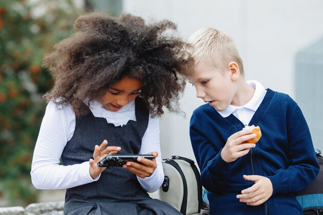 卷发一个非洲女孩和一个金发男孩坐在台阶上玩智能手机可爱人金发