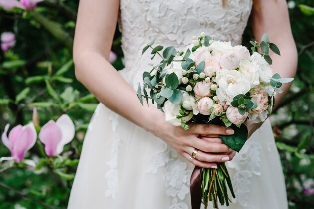 日期新娘手拿着一束美丽的婚礼花束 花束上有粉彩 粉色 白色的牡丹花 玫瑰花 绿叶 并在绿叶和木兰的衬托下用丝带装饰花女性礼服