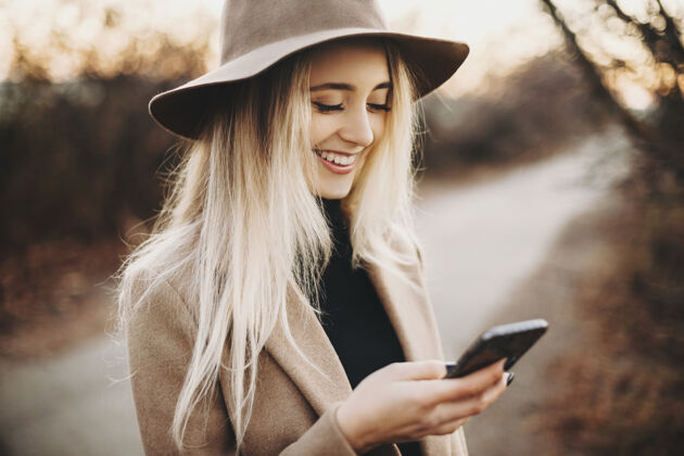 网络美丽的年轻女子 戴着优雅的帽子 站在秋日里愉快地微笑着使用智能手机乡村欢快在大自然中浏览智能手机的女士女性优雅和平