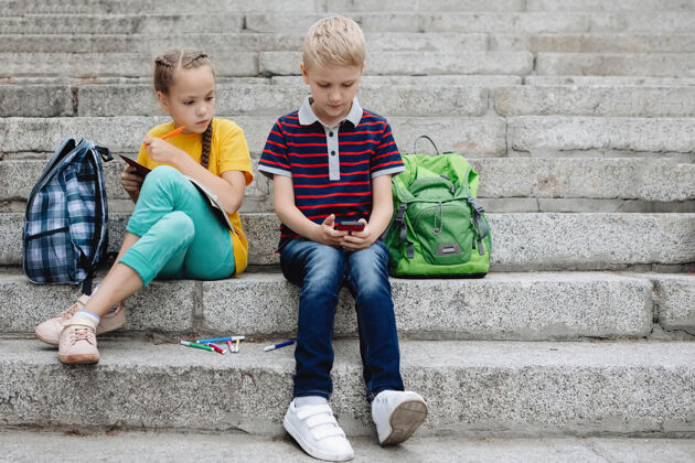 户外两个少年 一个男孩和一个女孩 手里拿着智能手机坐在台阶上两个颜色青少年