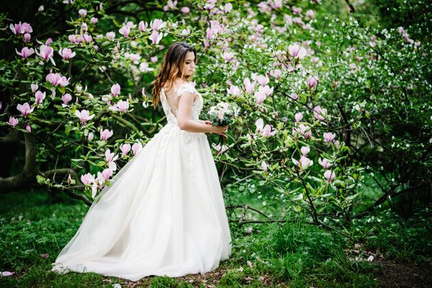 树新娘的肖像与婚礼花束站在背后 背景自然是紫色 粉红色的木兰花和绿色户外看向下完全长度阳光女性花园