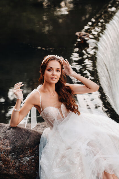 山河一位穿着白色礼服 戴着手套 赤脚的优雅新娘正坐在公园的瀑布旁 享受着大自然新娘女孩浪漫