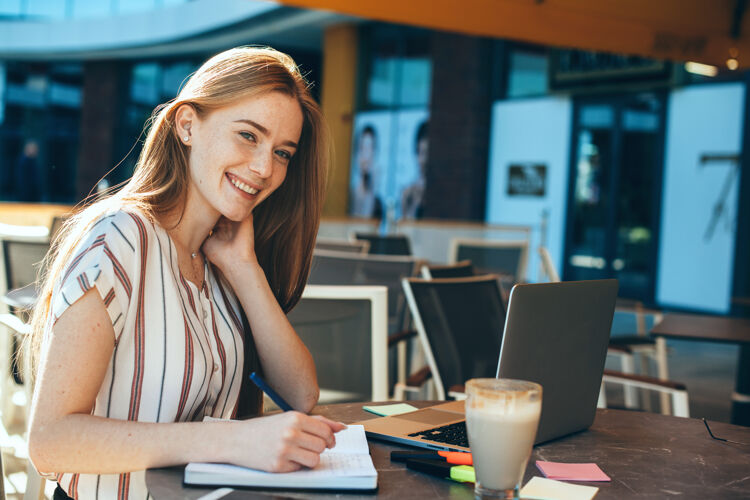 社交媒体一个可爱的年轻女商人在户外咖啡馆喝咖啡时写在笔记本上的肖像购买红色满头头发的女性微笑着咖啡馆自由职业者头像