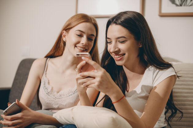 年轻两个可爱的白人女友坐在家里的沙发上看着智能手机屏幕大笑姐姐手机女人