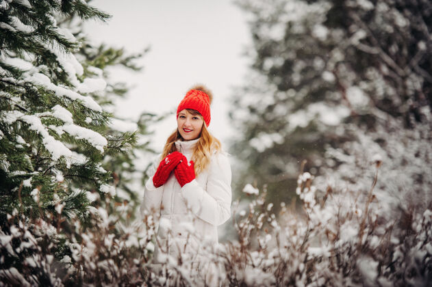 白帽子一个穿着白衣服 戴着红帽子的女人在寒冷的冬天的肖像森林女孩在冰雪覆盖的冬季森林里长美丽女人