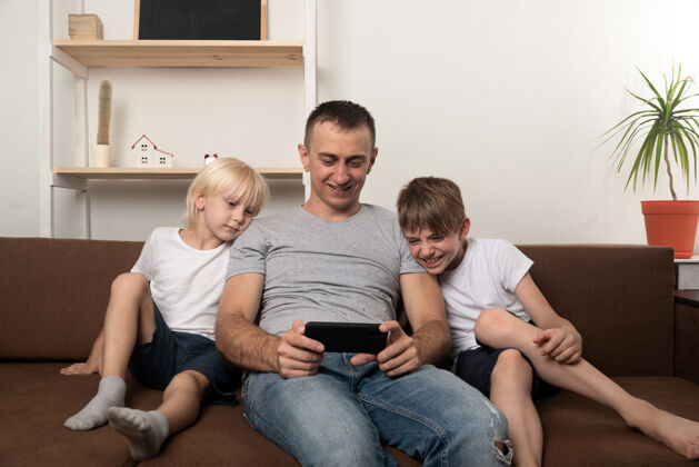 使用爸爸和两个儿子坐在沙发上看电视电话.家庭休闲哥哥爸爸室内