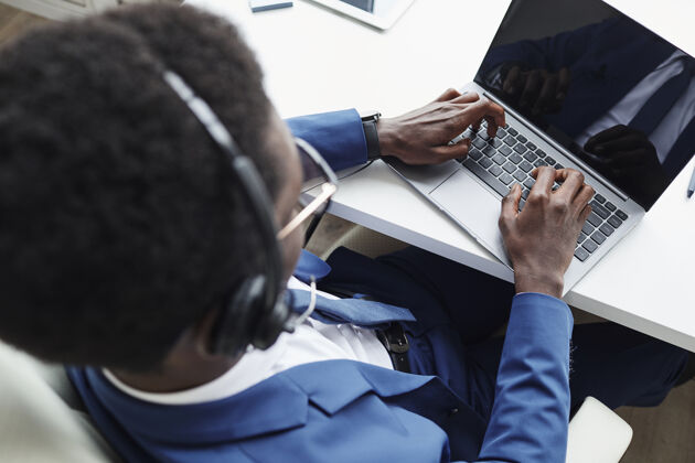 职业非洲商人戴着耳机在办公桌旁的笔记本电脑上打字的高视角照片他在网上交流白领技术打字
