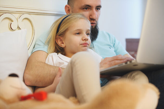 父母早上在家里 可爱的小女孩倚在爸爸的身上 看着他在床上的笔记本电脑屏幕男人学习电脑