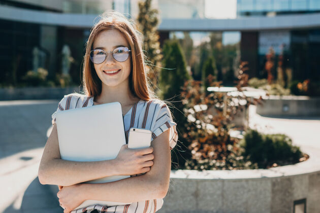 无线一位留着红头发和雀斑的美丽年轻女子的肖像 拿着笔记本电脑和智能手机对着现代化的办公室大笑女人肖像眼镜