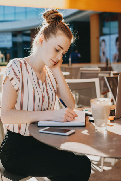 连接一个迷人的红发女人坐在外面的咖啡馆里写在笔记本上的侧视图打字数码笔记