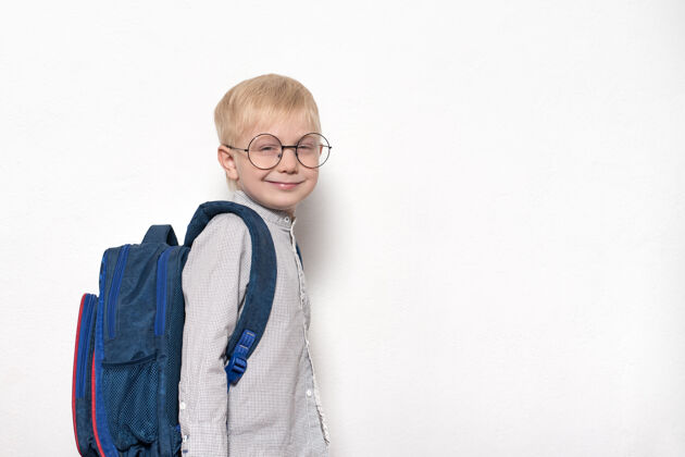 童年一个戴着眼镜的金发男孩的肖像 白色椅子上背着一个学校背包背景.学校概念学习学校表演