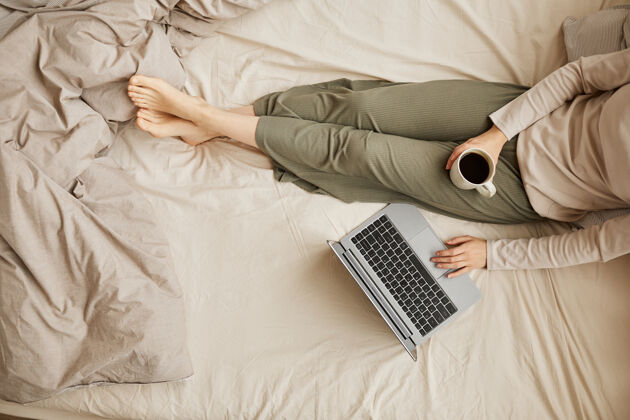 睡衣一个女人坐在床上喝着咖啡 早上用笔记本电脑的特写镜头家庭室内躺下床