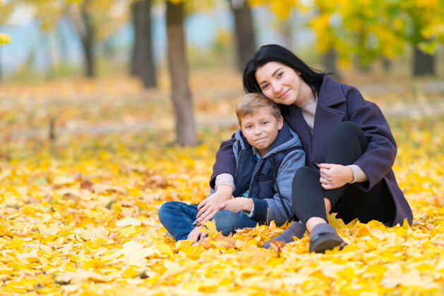 女人年轻女子抱着公园里十几岁的男孩坐在地上 满地都是秋天鲜艳的黄叶 看着他摄像头已满-长度与复制空间肖像树叶森林质量时间