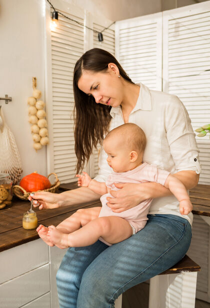 烹饪妈妈把女儿抱在怀里 用勺子在厨房的椅子上喂土豆泥女儿晚餐漂亮