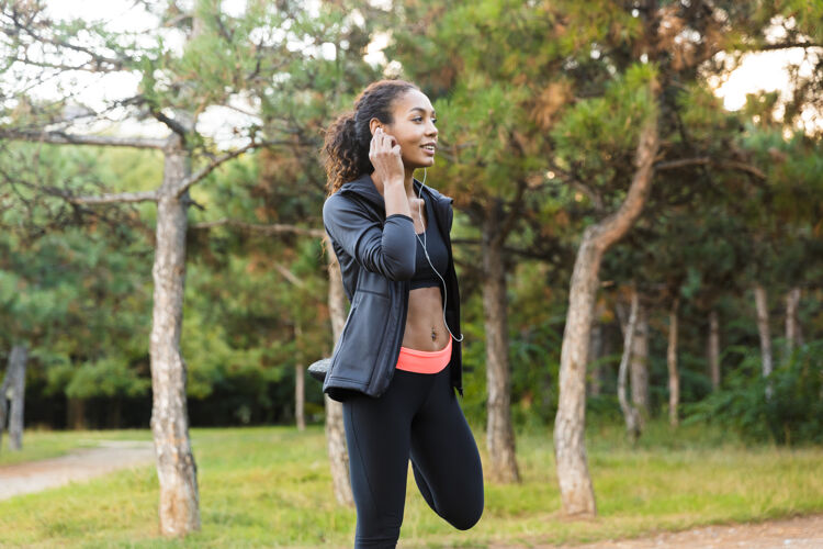 户外20多岁的非洲裔美国妇女穿着黑色运动服热身 在绿色公园伸展身体的画面女性听户外