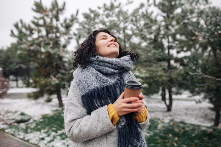公园穿着弗雷时装外套 戴着蓝色围巾的年轻女子站在那里喝着咖啡 准备去冬季的雪景公园温暖饮料独自