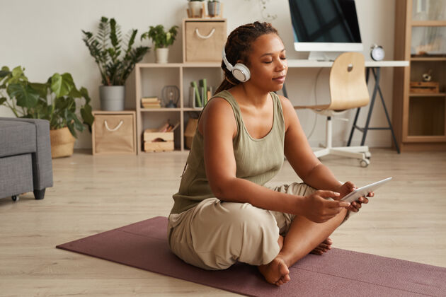 放松一位非洲妇女戴着无线耳机 闭着眼睛坐在运动垫上 用数字平板电脑听轻松的音乐运动垫家居内饰耳机