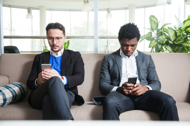 消息两个多元文化的男人坐在沙发上拿着手机短信通信网络