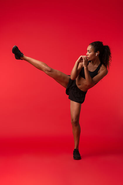 健身穿着黑色运动服的运动型非裔美国妇女在红墙外踢脚踢的肖像身体体操美国