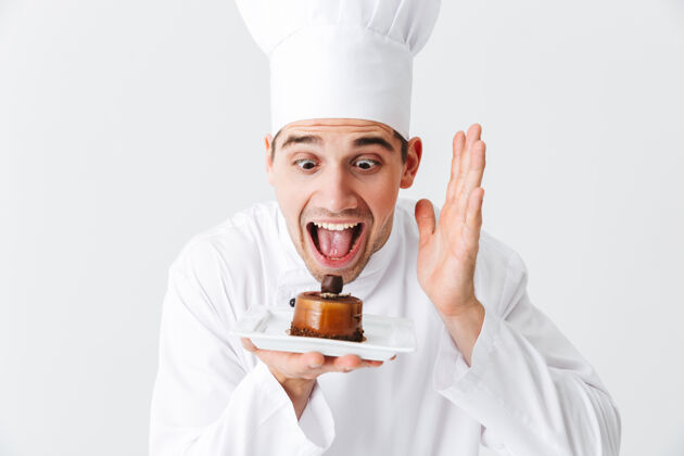 美食家开朗的厨师穿着制服 在白色的墙上展示一个盘子上的糕点厨房厨师面包房