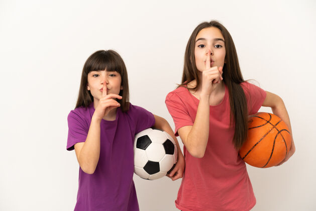 球在白色背景上孤立地踢足球和打篮球的小姐妹们表现出一种沉默的手势把手指放进嘴里孩子朋友手指